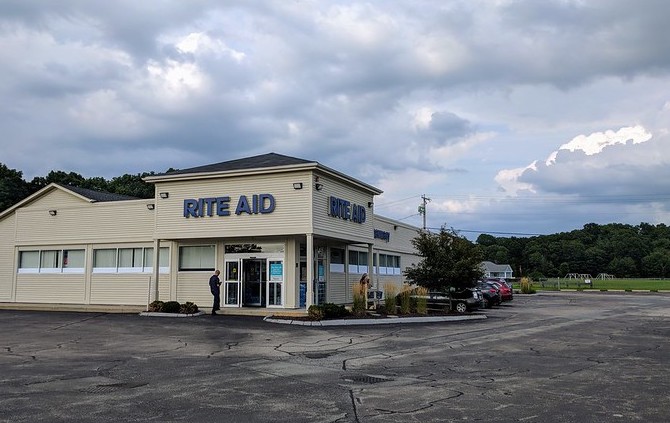 A tan building with shuttered windows and "Rite Aid" in blue letters above an open door sits behind an empty parking lot.