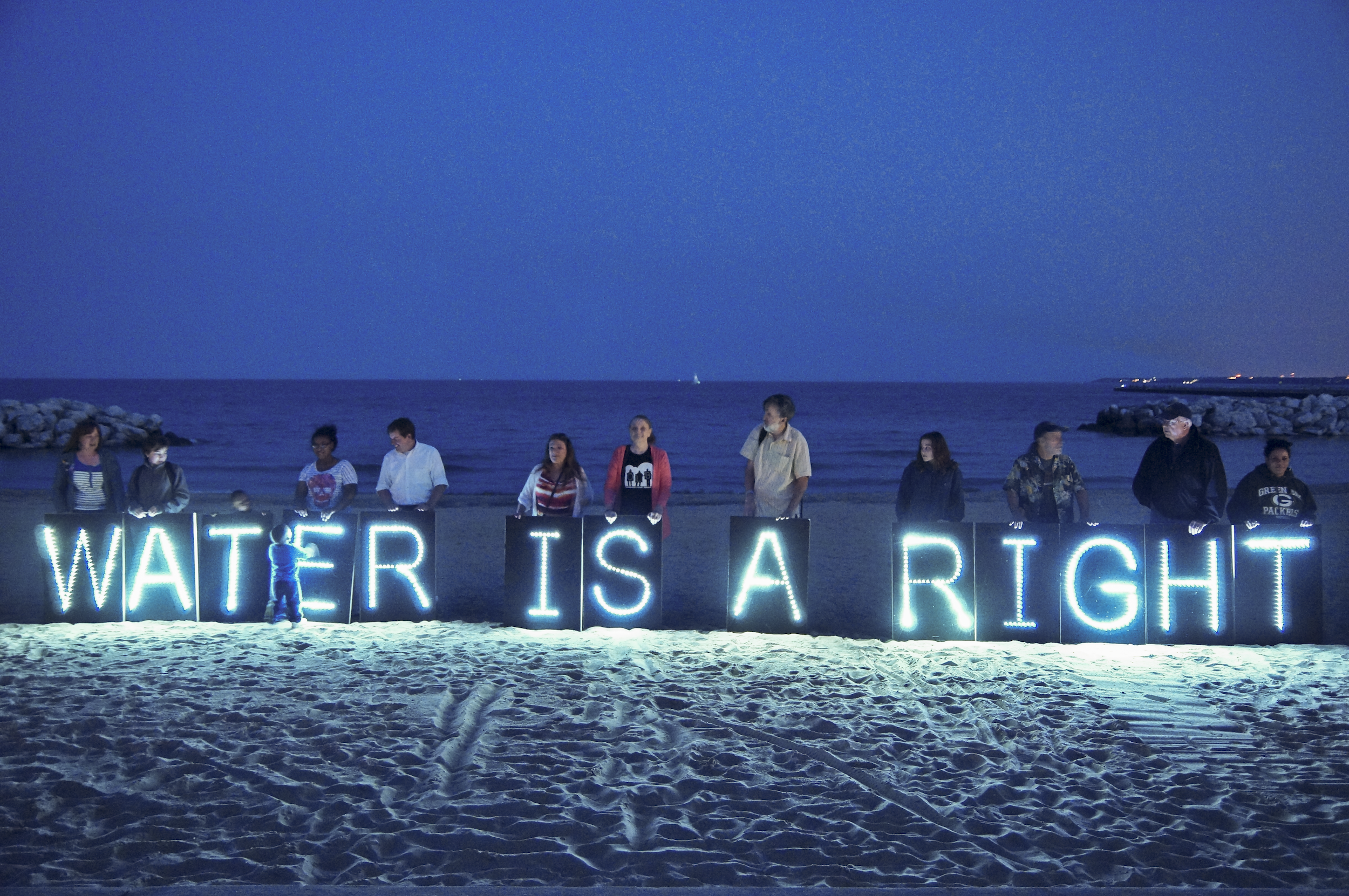 A small group of people standing in front of the ocean in moonlight holding up lit up signs that read "water is a right"