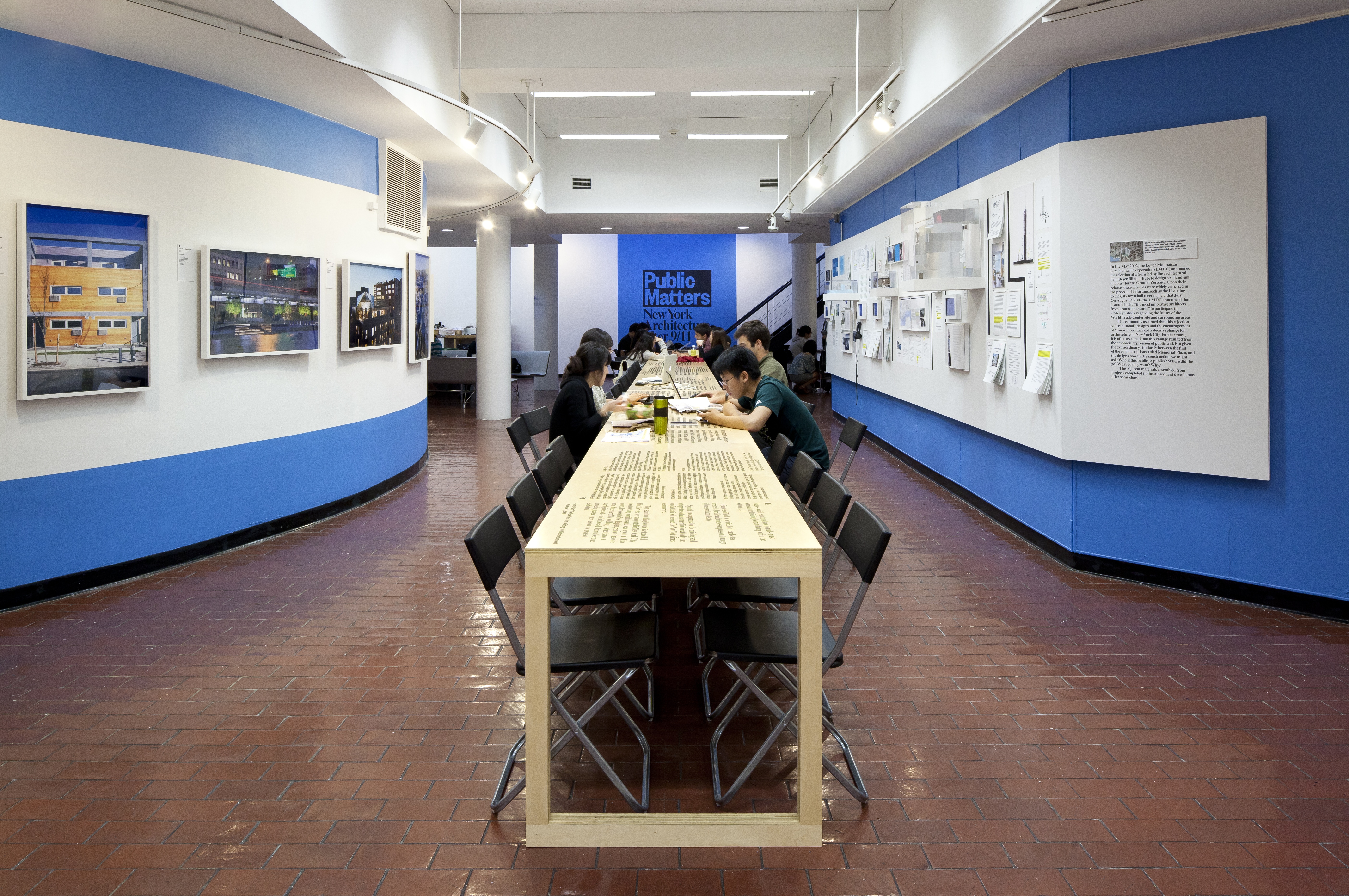 A long, narrow table with people working recedes into the background of an apparent cafe? gallery? On the left wall are framed photographs of buildings, on the right wall is a collection of paperwork, small screens, headphones, and small photographs. In the back, painted on the blue walls, is the black text "Public Matters: New York Architecture after 9/11." 
