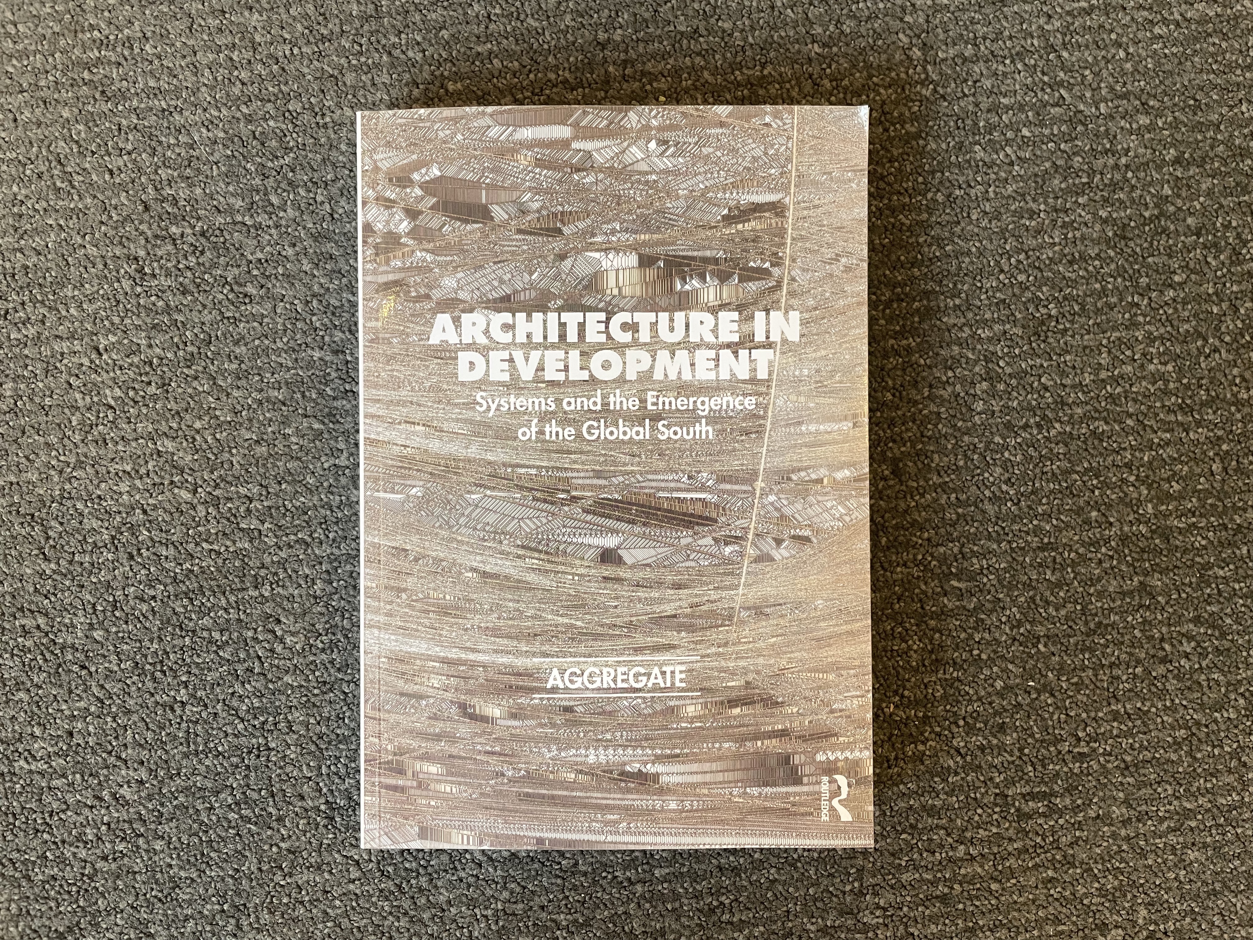 A book with a light brown and grey textured cover, potentially a line drawing or potentially photographic, reads "Architecture in Development: Systems and the Emergence of the Global South | Aggregate."