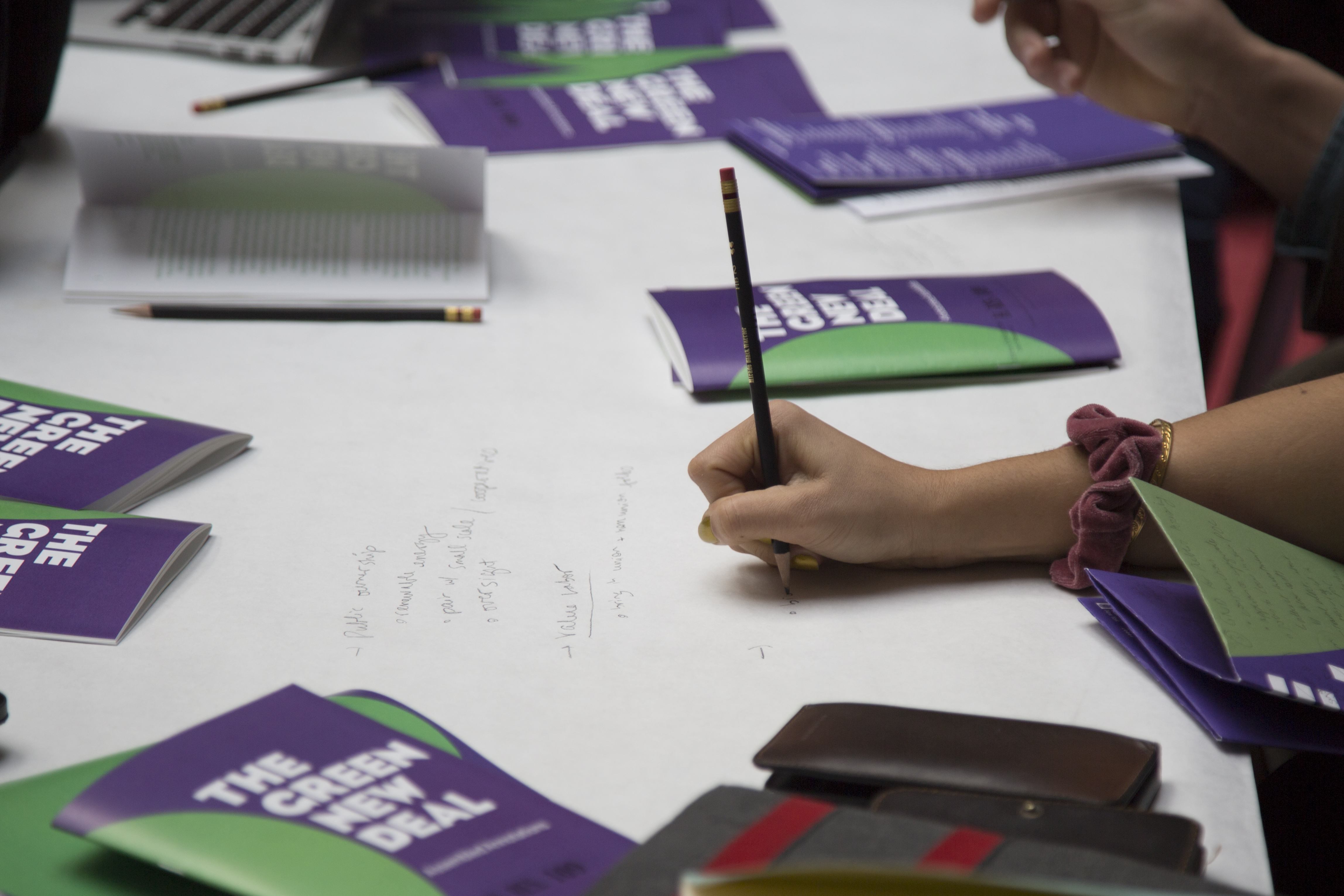A hand of a workshop participant holding a pencil, taking notes alongside scattered pamphlets titled, "The Green New Deal: Assembled Annotations."