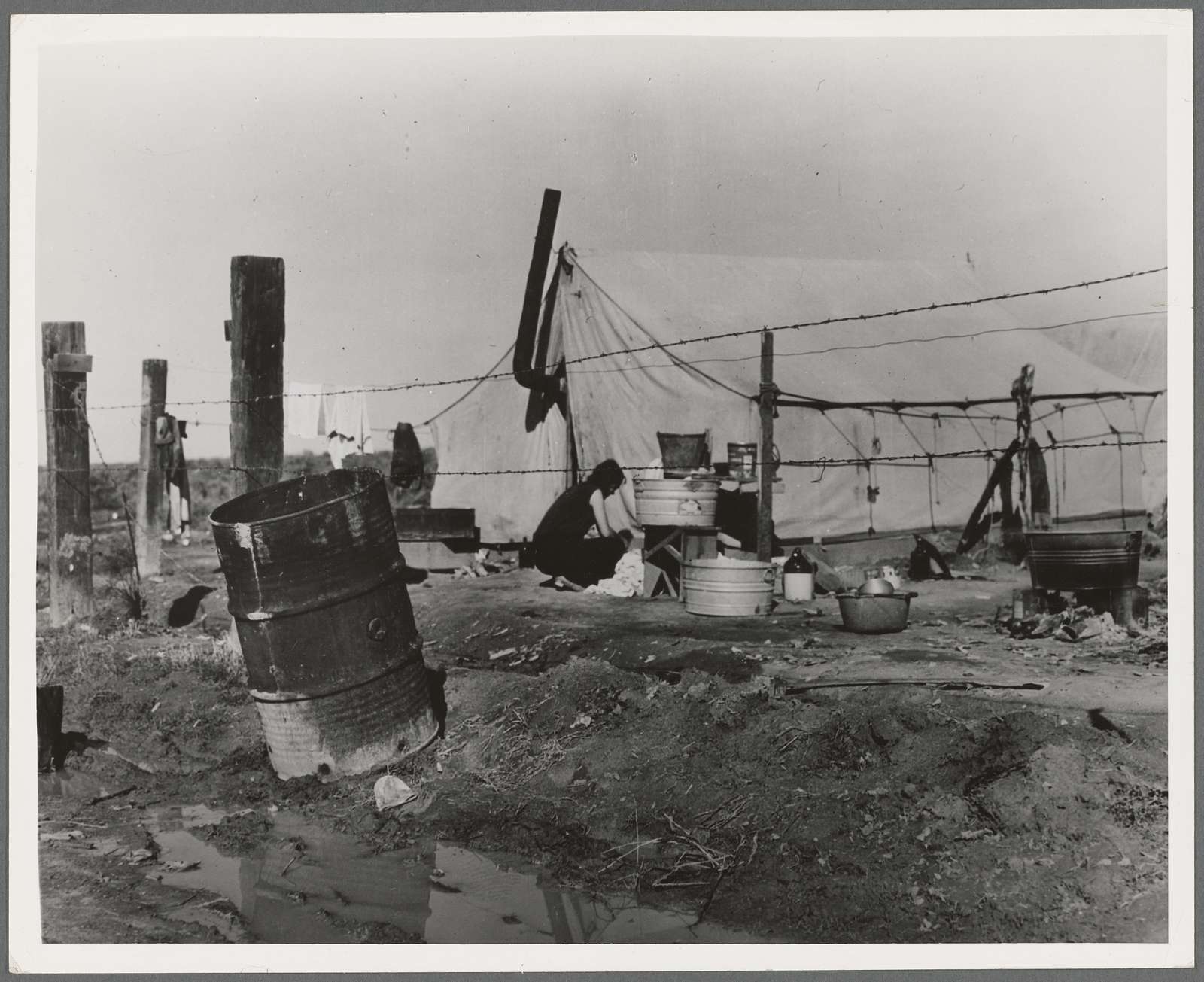 This black and white image shows canvas tent visible through a barbed-wire fence, with a crouched figure washing in the foreground.