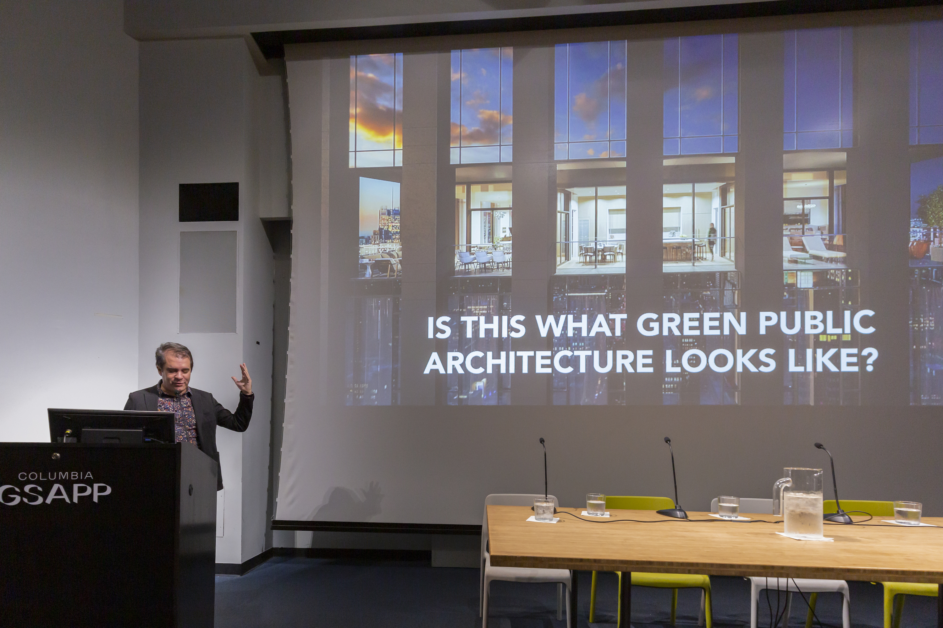 Andrés Jaque presenting at "Public Works for a Green New Deal." Jaque is seen standing behind a podium with his left arm raised. The slide on the auditorium screen reads "Is this what green public architecture looks like?"